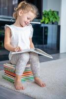 sonriente pequeño niña es sentado en apilar de para niños libros y hojeando mediante un libro con hada cuentos foto