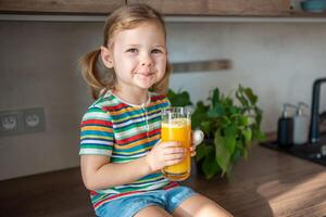 pequeño niña Bebiendo Fresco jugo sentado en mesa en hogar cocina foto