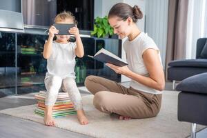 Little girl sits on a stack of children's books and watches cartoons on smartphone while her mother reads a book photo