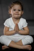 Tres años antiguo pequeño niña meditando en un loto actitud en un gris antecedentes en oscuro habitación foto