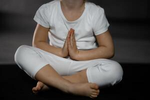 Tres años antiguo pequeño niña meditando en un loto actitud en un gris antecedentes en oscuro habitación foto