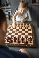 pequeño niña jugando ajedrez con su padre a el mesa en hogar cocina. el concepto temprano infancia desarrollo y educación. familia ocio, comunicación y recreación. foto
