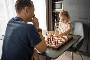 Father teaching his little daughter to play chess at the table in home kitchen. The concept early childhood development and education. Family leisure, communication and recreation. photo