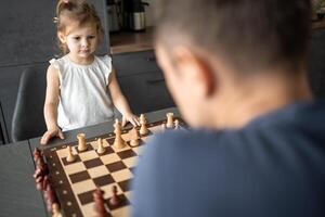 padre enseñando su pequeño hija a jugar ajedrez a el mesa en hogar cocina. el concepto temprano infancia desarrollo y educación. familia ocio, comunicación y recreación. foto