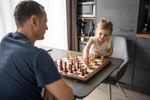 Little girl playing chess with her father at the table in home kitchen. The concept early childhood development and education. Family leisure, communication and recreation. photo