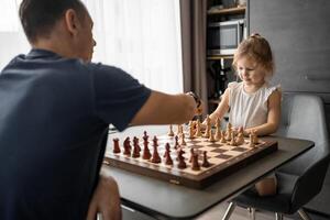 padre enseñando su pequeño hija a jugar ajedrez a el mesa en hogar cocina. el concepto temprano infancia desarrollo y educación. familia ocio, comunicación y recreación. foto