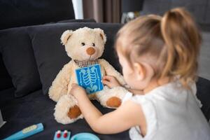 Little girl playing doctor with toys and teddy bear on the sofa in living room at home photo