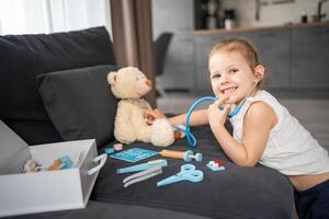 pequeño niña jugando médico con juguetes y osito de peluche oso en el sofá en vivo habitación a hogar foto