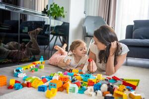 pequeño niña jugar con constructor juguete en piso en hogar con mamá o mujer niñera, educativo juego, familia a hogar gastar ocio ocupaciones hora juntos concepto foto