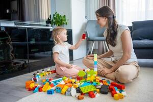 pequeño niña jugar con constructor juguete en piso en hogar con mamá o mujer niñera, educativo juego, familia a hogar gastar ocio ocupaciones hora juntos concepto foto