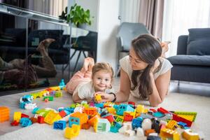 pequeño niña jugar con constructor juguete en piso en hogar con mamá o mujer niñera, educativo juego, familia a hogar gastar ocio ocupaciones hora juntos concepto foto