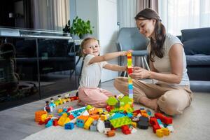 pequeño niña jugar con constructor juguete en piso en hogar con mamá o mujer niñera, educativo juego, familia a hogar gastar ocio ocupaciones hora juntos concepto foto