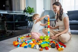 pequeño niña jugar con constructor juguete en piso en hogar con mamá o mujer niñera, educativo juego, familia a hogar gastar ocio ocupaciones hora juntos concepto foto