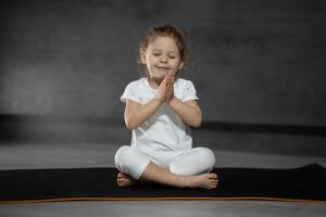 Tres años antiguo pequeño niña meditando en un loto actitud en un gris antecedentes en oscuro habitación. alto calidad foto