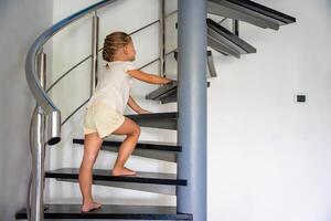 Little girl going up the stairs at home, child climbing spiral staircase. Dangerous situation at home. Child safety concept. photo