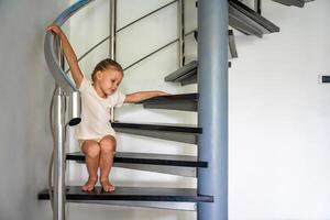 pequeño niña es sentado solo en espiral escalera a hogar. peligroso situación a hogar. niño la seguridad concepto. foto