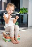 linda pequeño niña se sienta en apilar de para niños cuento de hadas libros y relojes dibujos animados en teléfono inteligente foto