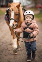 pequeño niña en protector chaqueta y casco con su marrón poni antes de montando lección. alto calidad foto