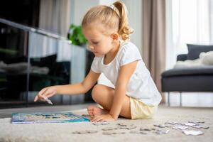 Little blonde girl sits at home on the carpet and collects puzzles photo