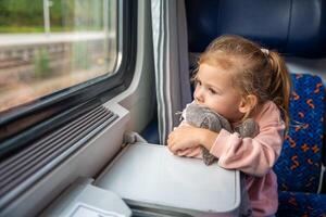 hermosa pequeño niña con juguete mirando fuera tren ventana afuera, mientras eso Moviente. de viaje por ferrocarril en Europa foto