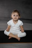 Tres años antiguo pequeño niña meditando en un loto actitud en un gris antecedentes en oscuro habitación. alto calidad foto