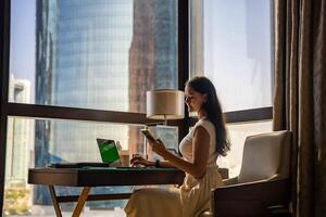 Stylish businesswoman freelancer working with laptop and smart phone, she is sitting on chair, enjoying panoramic view in city background. Low key photo. High quality photo