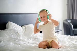 linda pequeño niña emocional disfrutando música utilizando verde niños auriculares en hogar cama. alto calidad foto