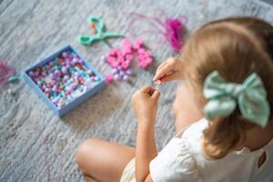 pequeño niña haciendo de madera rosario pulsera a hogar vivo habitación. para niños creatividad y el desarrollo de multa motor habilidades foto