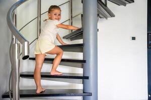 pequeño niña yendo arriba el escalera a moderno hogar, niño alpinismo espiral escalera. peligroso situación a hogar. niño la seguridad concepto. foto
