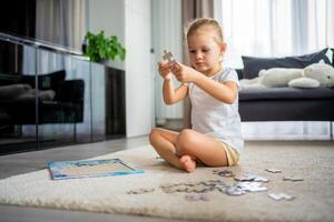 Little blonde girl sits at home on the carpet and collects puzzles photo