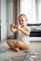 Little blonde girl sits at home on the carpet and collects puzzles photo