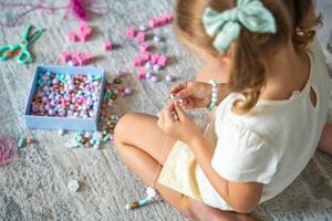 pequeño niña haciendo de madera rosario pulsera a hogar vivo habitación. para niños creatividad y el desarrollo de multa motor habilidades foto