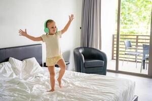 Little girl listening to the music with kids headphones and dancing on bed in happy morning. High quality photo