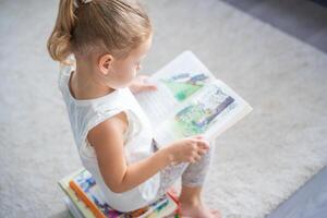 pequeño niña es sentado en apilar de para niños libros y hojeando mediante un libro con imágenes de hada cuentos foto