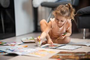 pequeño niña se sienta en el piso a hogar y sorteos con pinturas y cepillos en un colorante libro. temprano infancia creatividad y educación. alto calidad foto