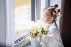 pequeño niña sentado por ventana con tulipán flores ramo. contento niño, adentro. de la madre día, San Valentín día o cumpleaños concepto. foto