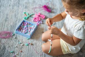 pequeño niña haciendo de madera rosario pulsera a hogar vivo habitación. para niños creatividad y el desarrollo de multa motor habilidades foto