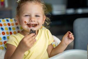 Little girl with blond hair eating homemade chocolate with dirty mouth and hands in home kitchen photo