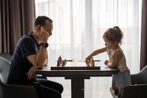 Father teaching his little daughter to play chess at the table in home kitchen. The concept early childhood development and education. Family leisure, communication and recreation. photo