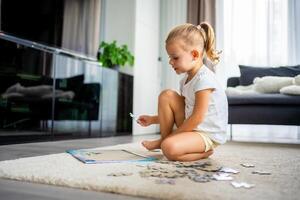 Little blonde girl sits at home on the carpet and collects puzzles photo