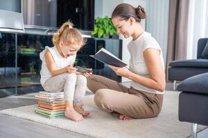 pequeño niña se sienta en un apilar de para niños libros y usos su teléfono inteligente mientras su madre lee un libro foto