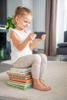 sonriente pequeño niña se sienta en apilar de para niños cuento de hadas libros y relojes dibujos animados en teléfono inteligente foto