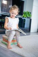 linda pequeño niña es sentado en apilar de para niños libros y hojeando mediante un libro con hada cuentos foto