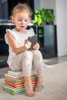 pequeño niña se sienta en un apilar de para niños cuento de hadas libros y obras de teatro con teléfono inteligente foto