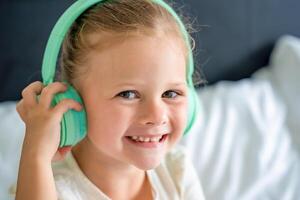 Smiling llittle girl enjoying music using green kids headphones in home bed. High quality photo