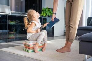 pequeño niña se sienta en un apilar de para niños libros con teléfono inteligente mientras madre da su un libro foto