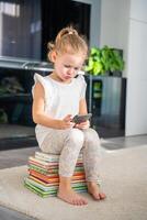 Little girl sits on stack of children's fairy-tale books and plays with her smartphone photo