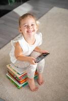 sonriente pequeño niña se sienta en apilar de para niños cuento de hadas libros con teléfono inteligente en su manos foto