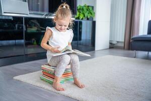 pequeño niña es sentado en un apilar de para niños libros y hojeando mediante un libro con hada cuentos foto