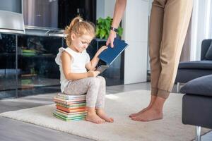 pequeño niña se sienta en apilar de para niños libros con teléfono inteligente mientras madre da su un libro foto